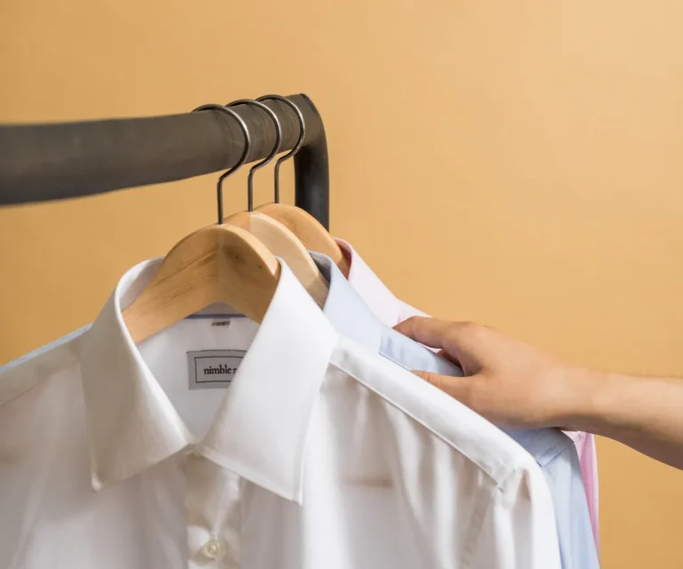 A hand selects a shirt from a rack holding three collared shirts on wooden hangers against a beige background.