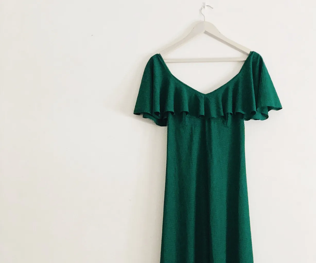 A green off-the-shoulder dress with ruffled sleeves hangs on a white hanger against a plain white wall.