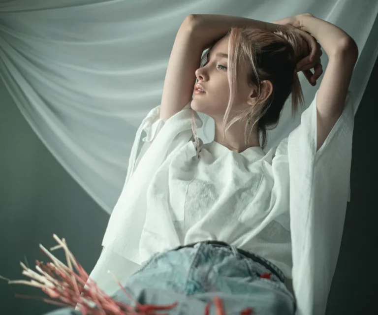 A woman with light hair poses with arms raised behind her head, wearing a white top and blue jeans, in front of a draped white fabric background.