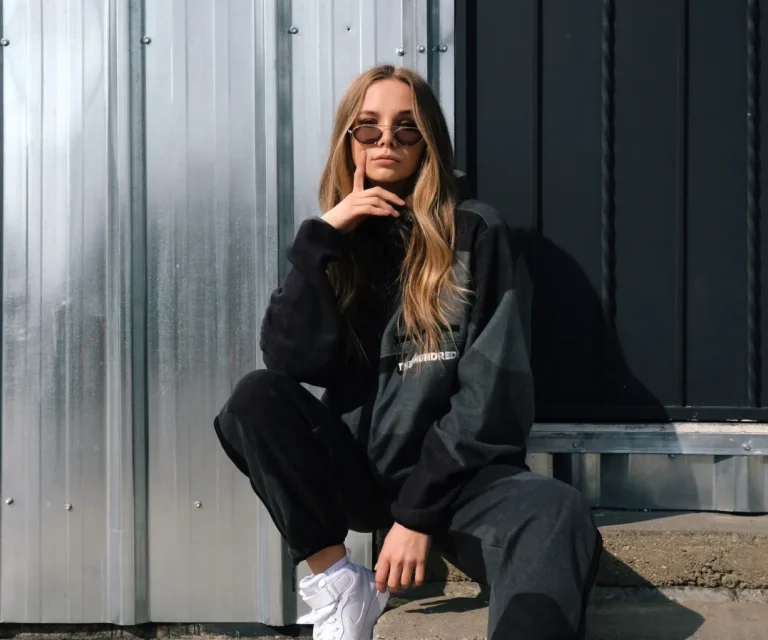 Woman wearing sunglasses and a dark tracksuit sits on a concrete step in front of a metal and black wall, resting her elbow on her knee and touching her face.