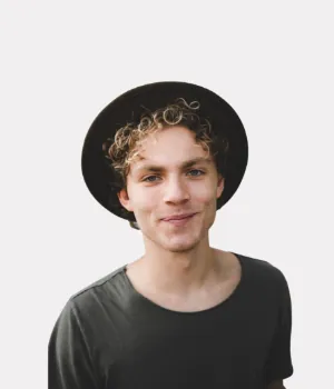 A young person with curly hair wearing a dark hat and a dark t-shirt, smiling at the camera against a plain white background.