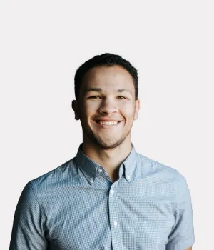 A young man with short dark hair wearing a light blue patterned button-up shirt smiles at the camera against a plain white background.