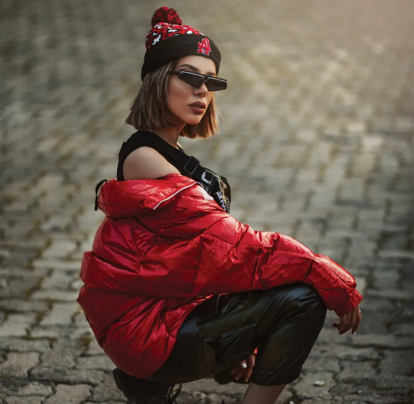 Woman squats on a cobblestone path wearing a red puffer jacket, black beanie, sunglasses, and black pants.